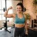 Mulher adulta com braços tonificados e definidos, sorrindo em um ambiente de treino em casa.